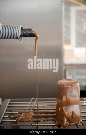 Un primo piano di cioccolato fuso che viene versato da una pipa in un contenitore in una fabbrica di cioccolato, mostrando la consistenza ricca e liscia. Germania Foto Stock
