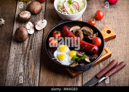 Frittura di uova di quaglia, salsicce, pomodori e funghi in padella in ghisa, prima colazione inglese in stile rustico Foto Stock