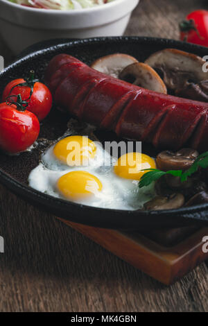 Frittura di uova di quaglia, salsicce, pomodori e funghi in padella in ghisa, prima colazione inglese in stile rustico Foto Stock