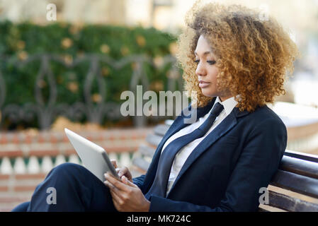 Bella black capelli ricci donna africana utilizzando computer tavoletta su un banco di URBAN. Imprenditrice indossare tuta con pantaloni e cravatta, acconciatura afro. Foto Stock