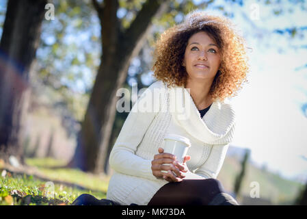 Bella giovane americano africano donna con acconciatura afro e gli occhi verdi indossano bianco inverno abito. Ragazza di bere il caffè nel parco seduto sull'erba abbiamo Foto Stock