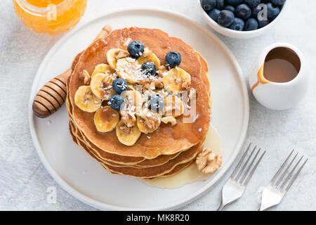 Pancake con mirtilli, banana, noci e miele sulla piastra bianca. Pila di gustosi pancake con sciroppo di caramello. Frittelle prima colazione con miele Foto Stock