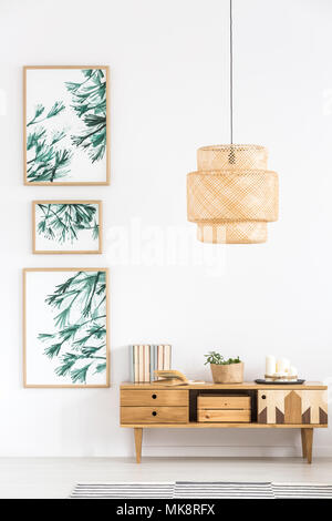 Lampada in rattan sopra armadio in legno contro il muro bianco con manifesti in modo semplice living room interior Foto Stock