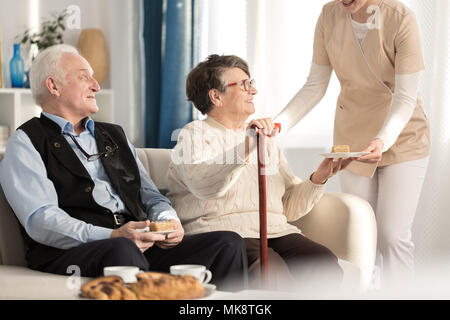 Geriatrico giovane con artrite seduto su un divano e di essere servito un pezzo di torta mentre è in attesa di un appuntamento dal dottore presso un privato di lusso clini Foto Stock