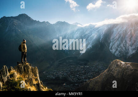 Giovani turisti in bright hat, pantaloni neri con uno zaino sorge su cliff's edge e guardando il misty Mountain Village e il ghiacciaio al sunrise, S Foto Stock