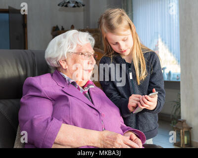 Un ragazzino di sette anni in azienda uno smartphone mostra qualcosa di divertente per il suo 95-anno-vecchia nonna. Lei è seduta su una sedia nel suo salotto e sono entrambi Foto Stock
