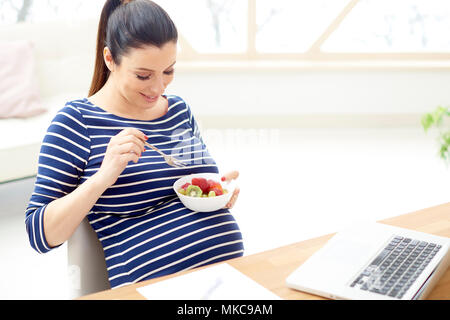 Bella giovane donna incinta mangiare una tazza di macedonia di frutta mentre è seduto nella parte anteriore del computer portatile e un momento di relax a casa. Foto Stock