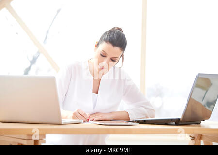 Ritratto di giovane sorridente assistente vendite donna facendo alcune pratiche mentre è seduto alla scrivania in ufficio nella parte anteriore del computer portatile e di lavoro on-line. Home office. Foto Stock