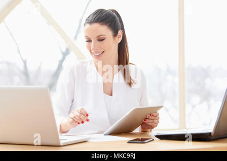 Ritratto di attraente giovane imprenditrice utilizzando tavoletta digitale e computer in ufficio. Foto Stock