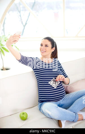 Felice donna incinta tenendo la sua mano 3d immagine ecografica e tenendo selfie comodamente seduti sul divano di casa. Foto Stock