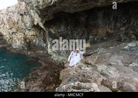 Giovane sposi sposa e lo sposo ride e sorrisi a ciascun altro, felice e gioioso momento. L uomo e la donna nel matrimonio abiti sedersi sulla roccia dello sfondo. Foto Stock