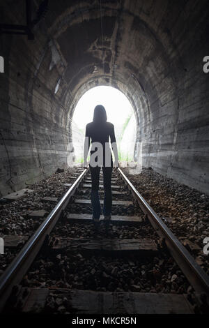 L'uomo profilarsi in un tunnel in piedi al centro dei binari ferroviari che guarda verso la luce alla fine del tunnel in una immagine concettuale Foto Stock