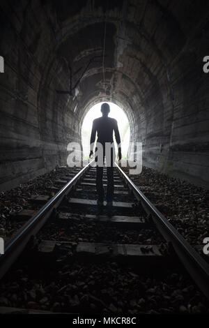 L'uomo profilarsi in un tunnel in piedi al centro dei binari ferroviari che guarda verso la luce alla fine del tunnel in una immagine concettuale Foto Stock