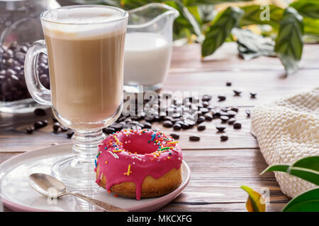 Tazza di caffè caldo latte bevande con vetrate a ciambella prima colazione la mattina presto sul tavolo in legno in stile rustico sparsi coffea grani e indoo verde Foto Stock