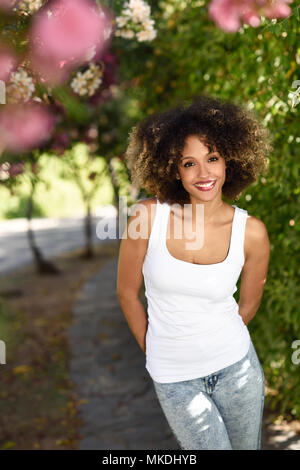 Giovane donna nera con acconciatura afro sorridente nel parco urbano. Ragazza misto bianco che indossa la t-shirt e jeans blu. Foto Stock