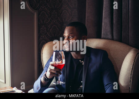 Pencive African American uomo in blu scuro giacca elegante guardando alla finestra e di bere il vino al ristorante Foto Stock