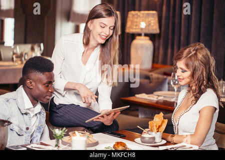 Gruppo di amici gustando il tempo in cafe insieme. Di carnagione scura guy e due bianchi affascinante Signore seduto a tavola a guardare le foto su tavoletta digitale e Foto Stock