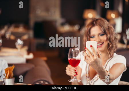 Felice giovane donna bianca con un bicchiere di vino prendendo selfie presso il ristorante Foto Stock