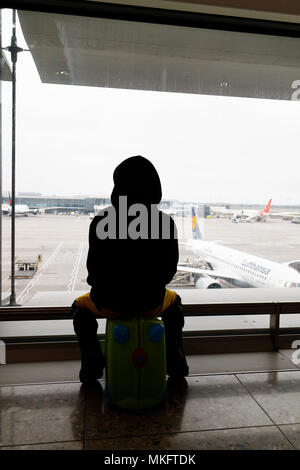 Un giovane ragazzo (5 yr old) seduto sulla sua valigia guardando i piani all'aeroporto di Londra Heathrow Foto Stock