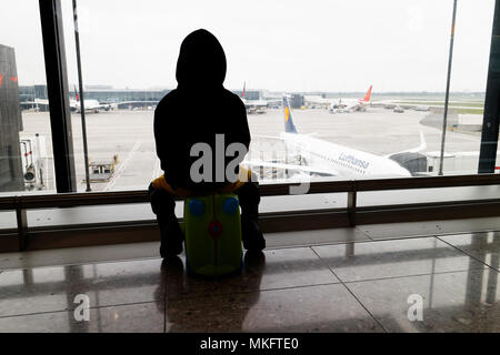 Un giovane ragazzo (5 yr old) seduto sulla sua valigia guardando i piani all'aeroporto di Londra Heathrow Foto Stock