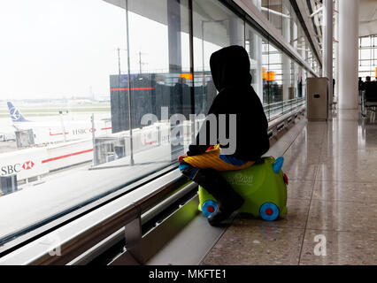 Un giovane ragazzo (5 yr old) seduto sulla sua valigia guardando i piani all'aeroporto di Londra Heathrow Foto Stock