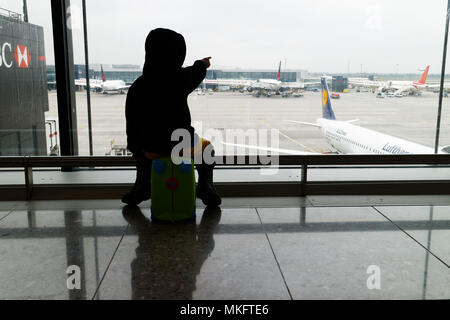 Un giovane ragazzo (5 yr old) seduto sulla sua valigia guardando i piani all'aeroporto di Londra Heathrow Foto Stock