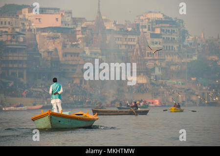 Un uomo è in barca a vela sulla sua barca di legno nel fiume sacro Gange nella città di Varanasi al tramonto. Sfocato Manikarnika ghat sullo sfondo. Foto Stock