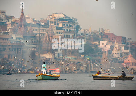 Un uomo è in barca a vela sulla sua barca di legno nel fiume sacro Gange nella città di Varanasi al tramonto. Sfocato Manikarnika ghat sullo sfondo. Foto Stock