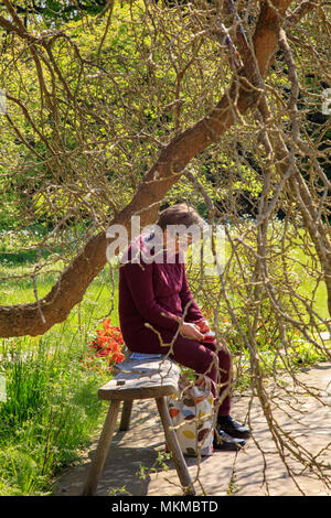 Una donna anziana si siede e poggia su una panca in legno sotto un gelso e utilizza un telefono mobile, Great Dixter, Northiam, East Sussex, Regno Unito Foto Stock