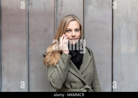 Giovane bella donna gentile indossando cappotto e scialle nero utilizza lo smartphone di fronte ad una parete metallica mentre guardando nella telecamera Foto Stock