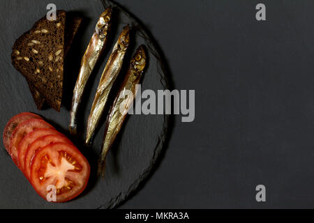 Capelin pesce del freddo affumicato sul round piastra di ardesia su sfondo nero. Possibilità di scrivere del testo. Foto Stock