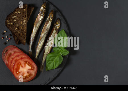 Capelin pesce del freddo affumicato sul round piastra di ardesia su sfondo nero. Possibilità di scrivere del testo. Foto Stock