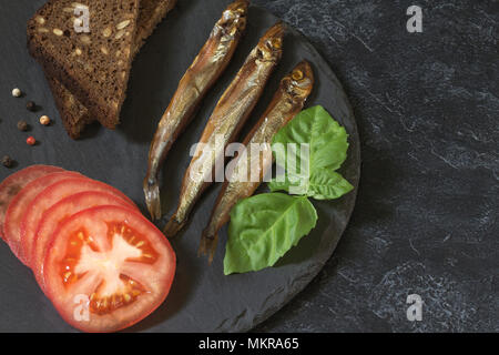 Capelin freddo affumicato pesce su un round piastra di ardesia su un nero-grigio sfondo irregolare. Possibilità di scrivere del testo. Foto Stock