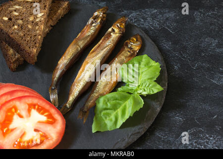 Capelin freddo affumicato pesce su un round piastra di ardesia su un nero-grigio sfondo irregolare. Possibilità di scrivere del testo. Foto Stock