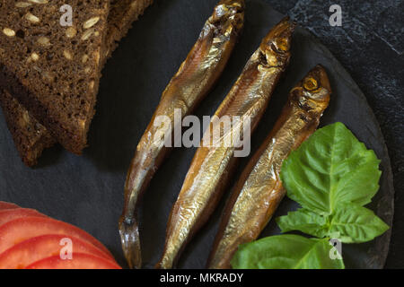 Capelin freddo affumicato pesce su un round piastra di ardesia su un nero-grigio sfondo irregolare. Possibilità di scrivere del testo. Foto Stock