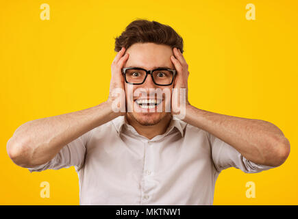 Eccitato giovane uomo in bicchieri tenendo le mani sul capo incredula guardando stupito dalla fotocamera su sfondo giallo. Foto Stock