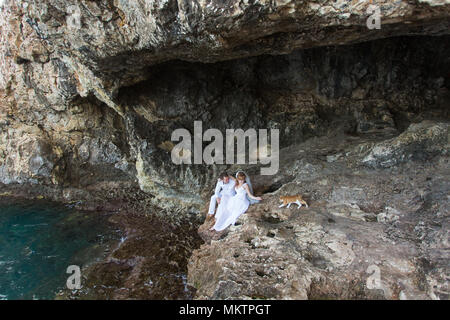 Giovane sposi sposa e lo sposo ride e sorrisi a ciascun altro, felice e gioioso momento. L uomo e la donna nel matrimonio abiti sedersi sulla roccia dello sfondo. Foto Stock