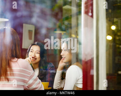 Tre felice giovane e belle donne asiatiche seduta a tavola in chat parlando in negozio di caffè o tè casa, shot attraverso il vetro di una finestra. Foto Stock