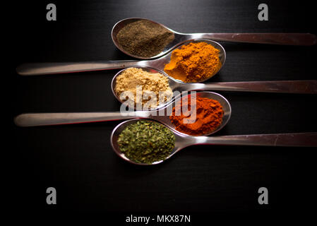 Spezie colorate in argento cucchiai, collocato su un tavoli in legno nero. Peperoncino, pepe, sale, basilico, senape Foto Stock