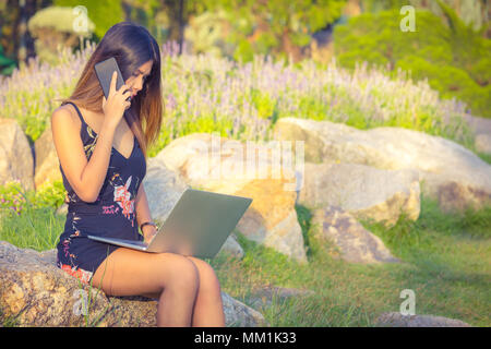 Attraenti donne business utilizzando il telefono e il computer portatile mentre a sedersi su una pietra nel parco durante il giorno Foto Stock