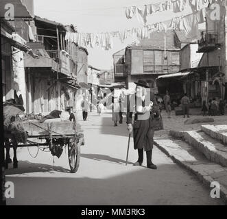 Degli anni Cinquanta, foto storiche di una strada sul retro della città vecchia di Gerusalemme, Israele mostra un asino e carrello e un anziano uomo ebraico con la barba lunga, in un cappotto, calze e hat a piedi con un bastone. Foto Stock