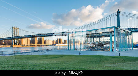 Manhattan Bridge al Ponte di Brooklyn Park. La parte inferiore di Manhattan è bagnata in inizio di mattina di sole e cielo blu e nuvole una serie di contrails erba verde. Foto Stock