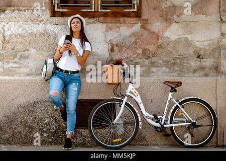 Felice pensieroso giovane donna sta godendo in estate il giorno soleggiato, appoggiata su un vecchio muro di casa, accanto alla moto con cesto fiorito. Foto Stock