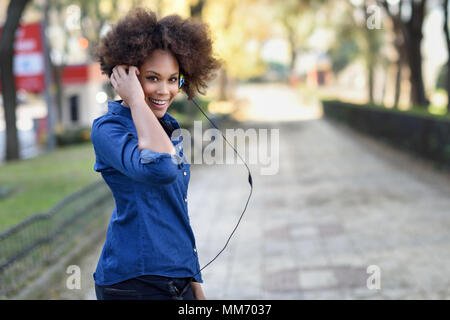 Giovane donna nera con acconciatura afro ascoltando la musica con le cuffie in background urbano. Funny misti donna che indossa maglietta blu e pantaloncini. Foto Stock