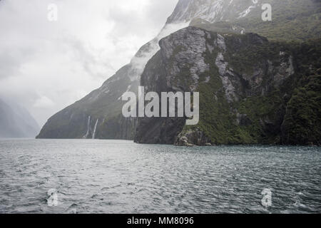 Cascate che scorre giù per la montagna rigogliosa e paesaggio di roccia nel Mare di Tasman ingresso del Parco Nazionale di Fiordland sotto un cielo di nebbia in Nuova Zelanda Foto Stock