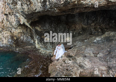 Giovane sposi sposa e lo sposo ride e sorrisi a ciascun altro, felice e gioioso momento. L uomo e la donna nel matrimonio abiti sedersi sulla roccia dello sfondo. Foto Stock