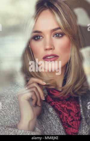 Considerato giovane donna bionda con gli occhi persi dalla finestra. Bella ragazza a casa indossando abiti invernali. Bella femmina con i capelli dritti e blu Foto Stock