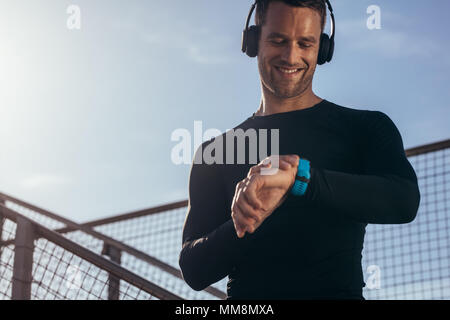 Uomo in piedi all'aperto con le cuffie utilizzando un smartwatch per monitorare i suoi progressi. Atleta maschio in appoggio e controllando la sua performance sul smartwa fitness Foto Stock