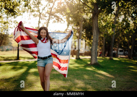 Bella giovane donna passeggiate nel parco e sventola bandiera americana. Ragazza sorridente con bandiera americana al parco. Foto Stock