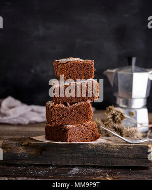 Una pila di pezzi quadrati di torta brownie su una tavola di legno vecchio cartone grigio, uno sfondo nero, uno spazio vuoto nella parte superiore Foto Stock